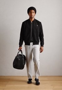 Black suede bomber jacket, gray trousers, black sneakers, holding a black duffel bag with white accents. Plain backdrop and wooden floor.