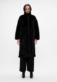 Abrigo negro de gran tamaño hecho de pelaje sintético texturizado, con un cuello alto, cierre de botones y mangas largas. Combinado con pantalones negros de pierna ancha.