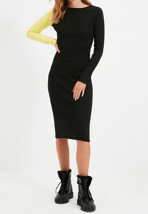 Femme portant une robe midi noire ajustée avec une manche jaune et des bottines à lacets noires, debout avec une main sur la hanche.