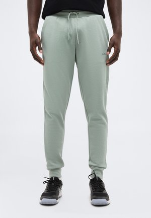 Mężczyzna w jasnych zielonych spodniach dresowych ze ściągaczem w pasie i czarnych butach sportowych, stojący z rękami swobodnie opuszczonymi wzdłuż ciała.