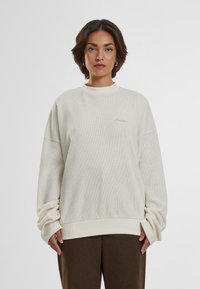 Helle beigefarbene Übergrößen-Sweatshirt aus strukturiertem Stoff mit geripptem Saum und Bündchen. Dezente gestickte Markenkennung auf der Brust.