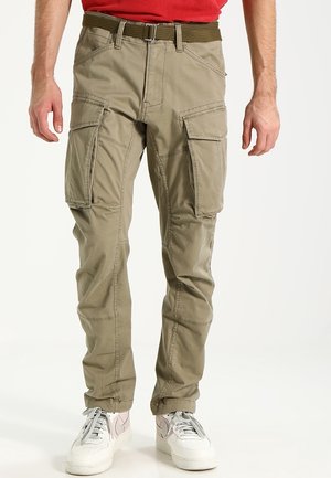 Homme portant un pantalon cargo beige avec poches à rabat, une ceinture marron, des baskets blanches et une chemise rouge, debout devant un fond uni.