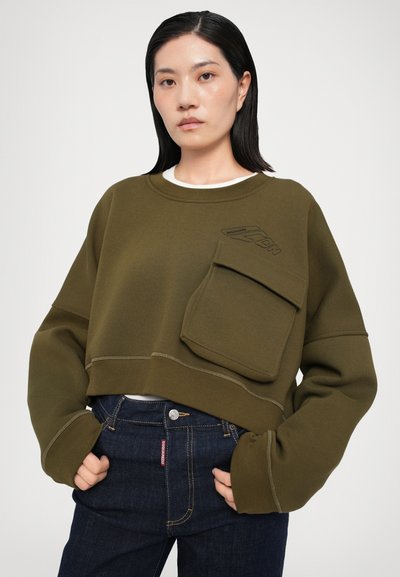 Tricou scurt de culoare verde măslin, dintr-un material moale, având o buzunar frontal cu clapete și cusături tonale. Asortat cu blugi negri.
