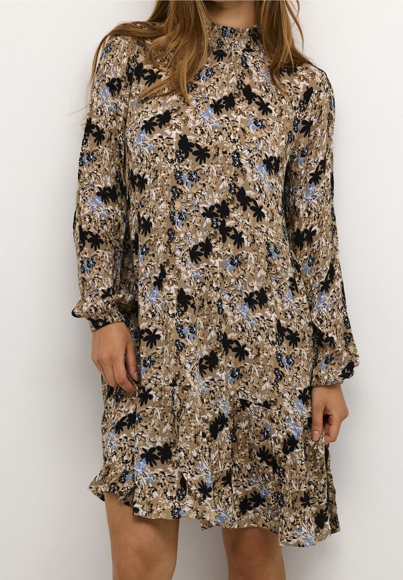 Femme portant une robe fleurie beige avec des fleurs noires et bleues, des manches longues bouffantes, un col haut et un ourlet à volants, debout contre un mur blanc.