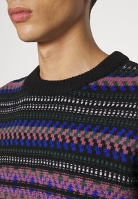 Neulepaita, jossa on pyöreä pääntie, monimutkaisia kuvioita mustassa, vaaleanpunaisessa, sinisessä ja vihreässä värissä, sekä ribbitekstuuri kauluksessa.