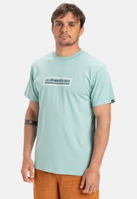 Světle modré tričko s krátkým rukávem vyrobené z bavlny, se černobílým logem s kostkovaným vzorem. Kulatý výstřih a volný střih.