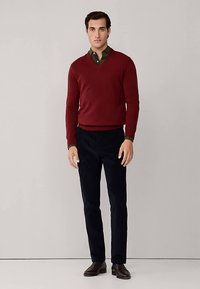 Bordeauxroter V-Ausschnitt-Pullover über einem dunklen grünen Hemd mit Kragen, kombiniert mit marineblauer Hose und braunen Lederschuhen. Einfaches, figurbetontes Design.