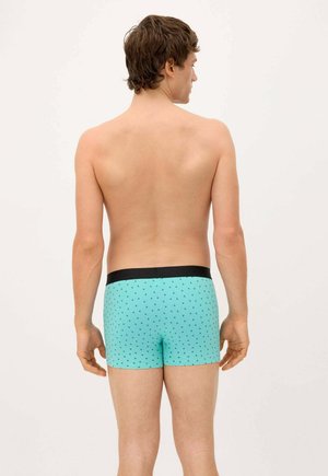 Joven de espaldas, vistiendo calzoncillos tipo bóxer azul claro con un pequeño estampado de palmeras negras y una cintura negra.