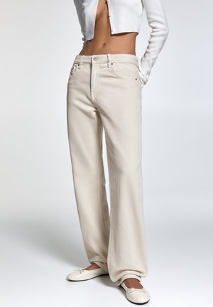 Kobieta ubranа w beżowe spodnie z szerokimi nogawkami z wysokim stanem, krótki biały sweterek i kremowe baleriny, stojąca na jednolitym tle.