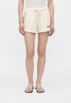 Kvinde iført cremefarvede snøre-shorts med multifarvede sidelommer og beige sandaler, stående foran en ensfarvet hvid baggrund.