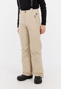 Beige skibroek met verstelbare bretels, ritszakken en een ontspannen pasvorm, gedragen met zwarte sneeuwlaarzen. Gladde textuur.