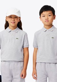 Två barn som bär gråa polotröjor i bomull, med korta ärmar och en liten grön logo. Ett barn har en vit keps.