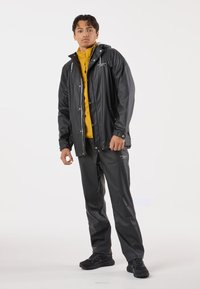 Wodoodporna czarna kurtka z kapturem i zapięciem na zamek, nałożona na żółtą bluzę zapinaną na zamek. Czarnie spodnie i trampki dopełniają strój.