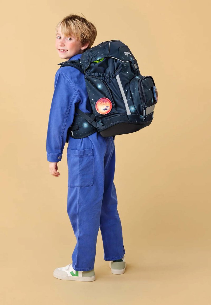 Jeune garçon en bleu de travail portant un sac à dos noir sur le thème de l'espace avec des patchs de planètes, debout et regardant par-dessus son épaule.
