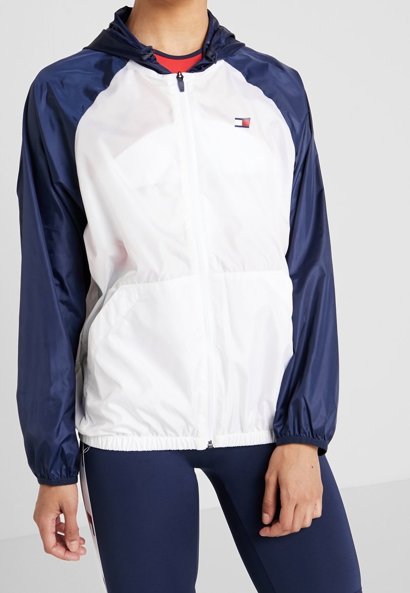Giacca a vento leggera con cappuccio, bianca e blu navy, dotata di zip frontale, polsini elasticizzati e un piccolo logo sul petto.