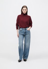 Maglione a collo alto color bordeaux, vestibilità comoda; jeans azzurri a gamba larga con dettagli usurati; ballerine nere; sfondo bianco semplice.