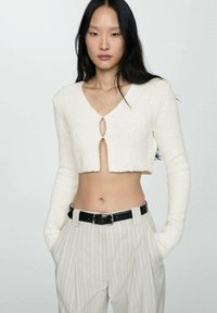 Pulover alb tricotat, scurt, cu mâneci lungi și detaliu tip clapă în față, asociat cu pantaloni deschiși la culoare cu dungi și o curea neagră.