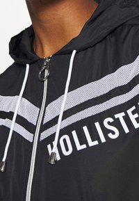 Mustat tuulitakit, joissa on huppu, sisältävät valkoisia verkkojuovia, edessä vetoketjun, jossa on pyöreä vetoketjunveto, ja "HOLLISTER" on painettu valkoisella.