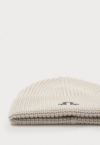 Knit beanie in off-white with vertical ribbing. Features a turned-up cuff and a small black logo accent. Soft texture and fitted shape.