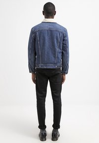 Džínová bunda tmavě modré barvy s bílým ovčím límcem, stylizovaná s černými slim-fit kalhotami a černými botami, z pohledu zezadu.