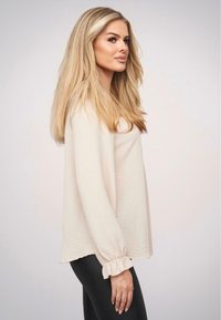 Maglietta lunga color beige chiaro con tessuto testurizzato e polsini arricciati, abbinata a leggings neri, mostrata in vista laterale su uno sfondo neutro.