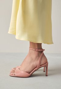 Sandales à talons hauts en cuir rose clair avec une bride autour de la cheville et un bout ouvert, portées avec une robe jaune fluide, sur une surface grise.