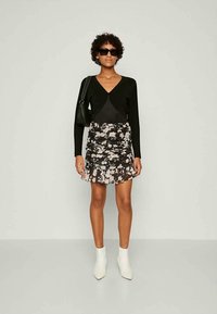 Femme portant un cardigan noir, une mini-jupe floral froncée, des bottines blanches, un sac à main noir et des lunettes de soleil sombres, se tenant devant un fond uni.