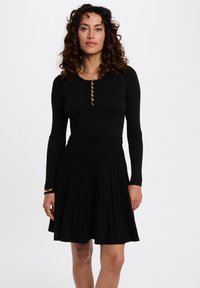 Schwarzes, geripptes Kleid mit langen Ärmeln, tailliertem Oberteil, knopffixiertem Ausschnitt und plissiertem Rock, der eine strukturierte Oberfläche aufweist.