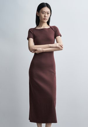 Žena s dlhými čiernymi vlasmi, oblečená v priliehavé hnedé midi šaty s krátkym rukávom, stojaca so založenými rukami pred jednoduchým svetlošedým pozadím.