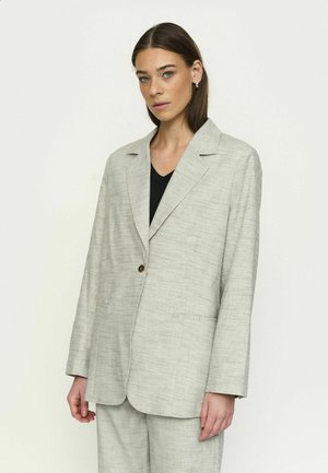 Sirged pruunide juustega naine kannab heledat halli tekstuuriga ülikonnajakki ja sobivaid pükse musta pluusi kohal, poseerides valgel taustal.