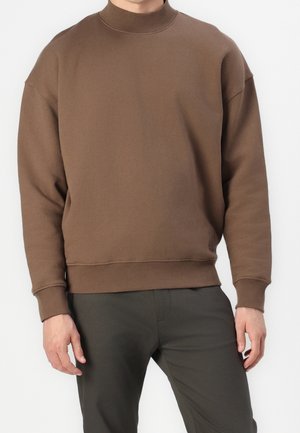 Mann trägt ein schlichtes braunes Sweatshirt mit hohem Kragen und dunkelgrüne Hose, steht vor weißem Hintergrund.