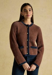 Brūns, pūkains cardigan ar tumši zilu apmali, pogām un kabatām. Garas piedurknes un apaļa kakla izgriezums, ar teksturētu apdari.