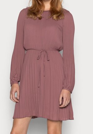 Kobieta ubrana w fioletową plisowaną sukienkę z długim rękawem i wiązanym paskiem w talii, stojąca na tle jednolitego, jasnego tła.