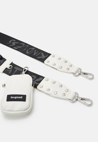 Sangle en tissu blanc et noir avec des fermoirs métalliques, des accents en cuir blanc à clous, et une petite pochette blanche zippée portant l'étiquette "Desigual".