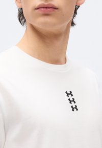 Νεαρό άτομο που φορά άσπρο πουκάμισο με τρία μαύρα λογότυπα Under Armour τοποθετημένα κατακόρυφα στο στήθος.