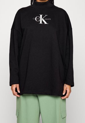 Person iført en løs sort langærmet top med Calvin Klein Jeans-logo og lysegrønne cargobukser.