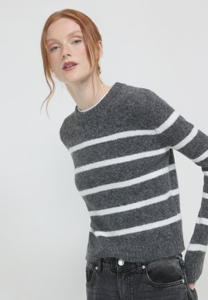 Femme aux cheveux roux portant un pull en peluche gris avec des rayures horizontales blanches et un jean noir, devant un fond clair uni.