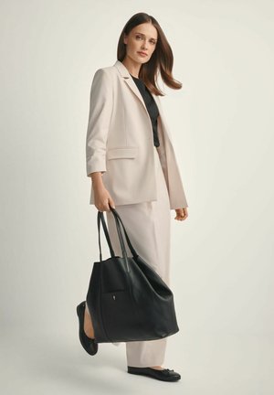 Kobieta w jasnobeżowym garniturze i czarnym topie niesie dużą czarną skórzaną torbę na ramię, idąc na tle jasnego tła.