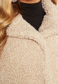 Beige tekstureret frakke i blødt, krøllet fleece-materiale, med en stor ombukket krave og klare knapper. Båret over en sort turtleneck.