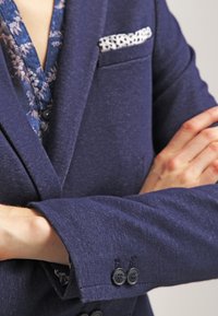 Marynarka w kolorze granatowym wykonana z teksturowanej tkaniny, z klapą z wycięciem, dwoma guzikami na mankiecie oraz wzorzystą poszetką schowaną w piersiowej kieszeni.