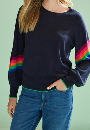 Femme portant un pull bleu marine avec des rayures arc-en-ciel sur les manches et une bordure verte, associée à un jean bleu, main dans la poche sur un fond clair.