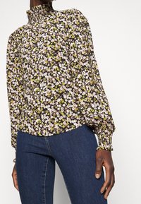 Bloemmotief blouse met hoge hals, pofmouwen en geplooide manchetten; kleuren zijn roze, geel en zwart op een lichtgewicht stof.