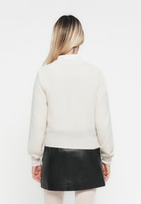 Vrouw met lang blond haar, gekleed in een witte trui en een zwarte leren rok, staande met haar rug naar de camera tegen een witte achtergrond.