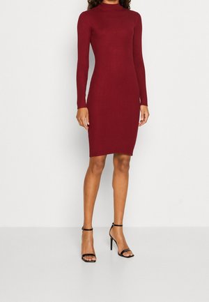 Vrouw draagt een aangesloten, lange mouwen, geribde bordeauxrode jurk tot op de knie met zwarte enkellaarsjes met hoge hak en enkelbandje, staand.