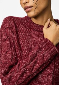 Burgundijas krāsas adīta džemperis ar cilpu adījuma rakstu, ribotu apļa kaklu un teksturētām piedurknēm. Mīksta, bieza dzija.