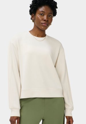 Vrouw met kort krullend haar, die een crèmewit shirt met lange mouwen en olijfgroene broek draagt en licht opzij kijkt.