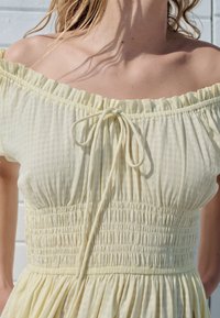 Gul axelbandslös klänning med smockad överdel, texturerat tyg och knytning vid halslinningen; lätt och flödande design.