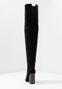 Botas negras de ante por encima de la rodilla, con un tacón grueso y un diseño partido en la parte superior. Detalle de cremallera en el lado.