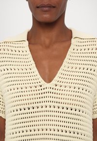 Personne portant un haut en tricot beige à manches courtes avec un col en V et un motif ajouré laissant apparaître la peau en dessous.