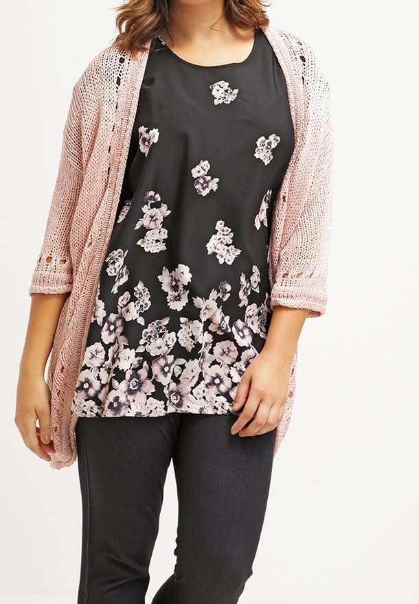 Femme portant un haut noir à motifs floraux, un cardigan en maille rose clair à manches trois-quarts, et un pantalon ajusté foncé, debout devant un fond blanc.